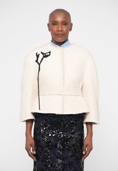 Cremfarbener Wolljacke mit Peplum-Design, ausgestattet mit einer schwarzen floralen Stickerei auf der linken Seite. Kombiniert mit einem marineblauen, mit Pailletten besetzten Rock.