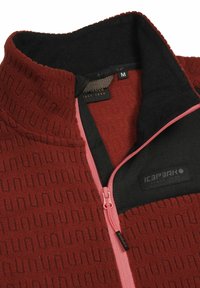 Giacca in pile rossa con un motivo testurizzato, accenti neri e rosa, colletto alto e zip frontale. L'etichetta indica la taglia M.