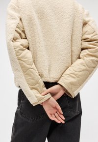 Personne portant une veste sherpa crème avec des manches matelassées, les mains jointes dans le dos, et un jean noir sur un fond uni.