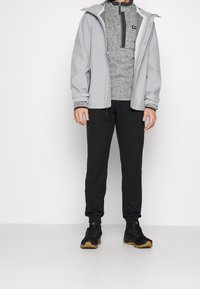 Giacca impermeabile grigia, maglione grigio con colletto alto, pantaloni neri e scarpe nere con suole beige, esposti su uno sfondo bianco.