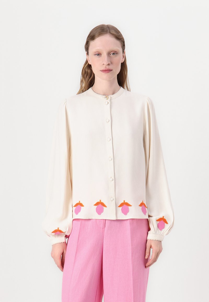 Cremefarbener Blusen mit langen, gepufften Ärmeln, Knopfverschluss und floralem Stickmuster in Pink und Orange am Saum. Kombiniert mit rosa Hosen.