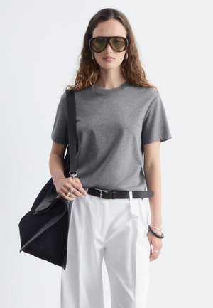Femme aux longs cheveux ondulés porte de grandes lunettes de soleil teintées, un t-shirt gris, un pantalon blanc, une ceinture noire, des bijoux en argent, et porte un sac à bandoulière noir.