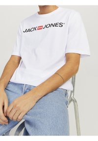 Camisa de algodón blanca con un atrevido logo negro de "JACK & JONES". Corte casual con mangas cortas, combinada con unos jeans de mezclilla azul claro.