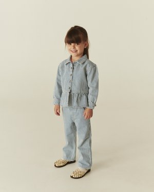 Young girl smiling, wearing blue and white striped jacket and pants, standing on a neutral background with beige woven shoes.