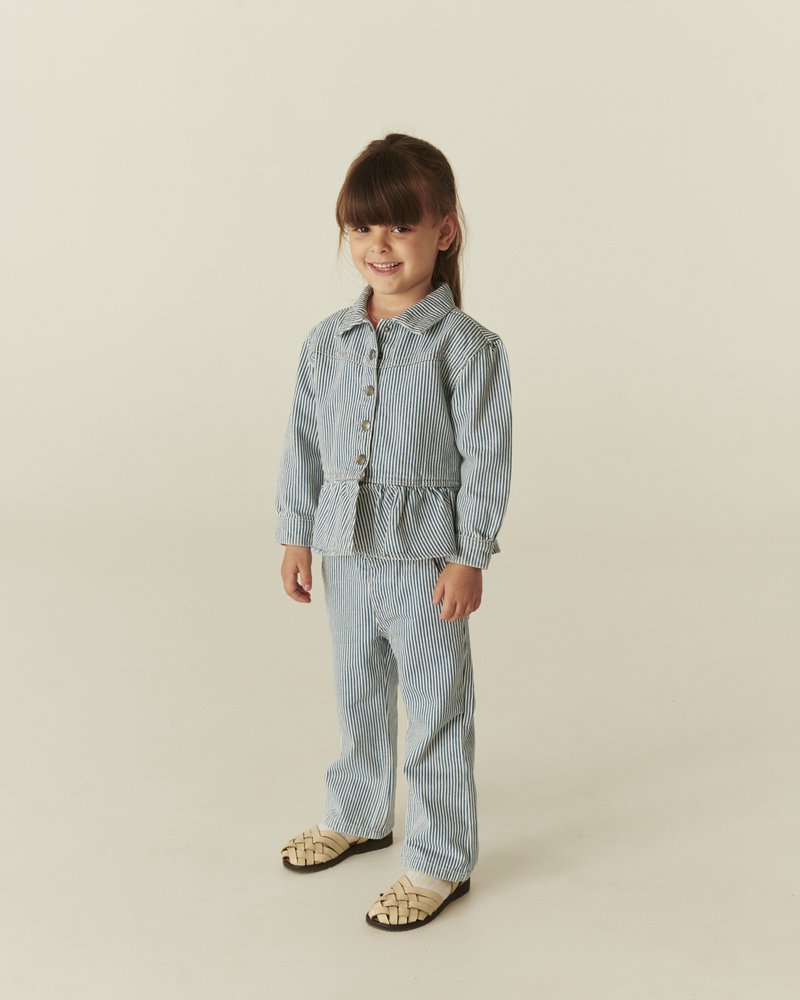 Νεαρή κοπέλα που χαμογελά, ντυμένη με μπλε και λευκή ριγέ ζακέτα και παντελόνι, στέκεται σε ουδέτερο φόντο με μπεζ πλεκτά παπούτσια.