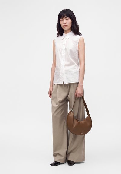 Femme aux cheveux noirs portant une chemise blanche sans manches, un pantalon beige à jambes larges, des chaussures noires, tenant un sac à main marron en forme de croissant sur fond blanc.
