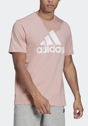 Mand iført lyserød Adidas t-shirt med stort hvidt logo og lysegrå Adidas shorts, stående mod ensfarvet baggrund.