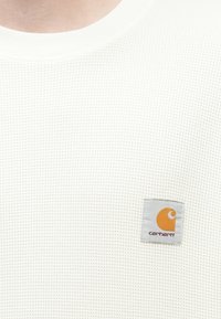 Λευκό πουλόβερ με υφή βάφλας και στρογγυλή λαιμόκοψη, με γκρι τμήμα που φέρει πορτοκαλί λογότυπο. Το υλικό έχει υφή που βελτιώνει την αναπνοή.