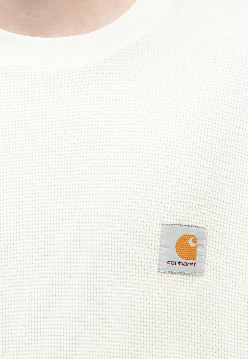 White waffle-knit crewneck sweater featuring a gray patch with an orange logo. Material has a textured appearance, enhancing breathability.