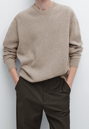 Personne portant un pull beige à col rond et manches longues légèrement rentré dans un pantalon foncé, les mains dans les poches.