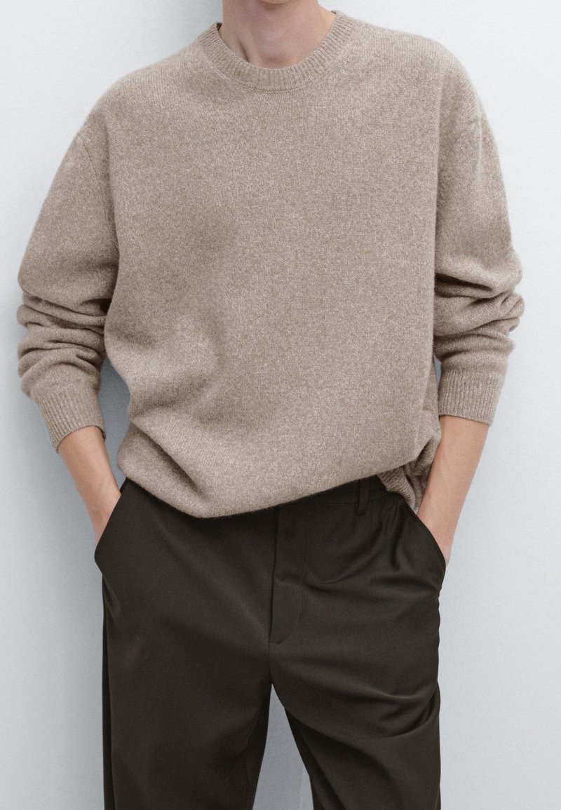 Personne portant un pull beige à col rond et manches longues légèrement rentré dans un pantalon foncé, les mains dans les poches.