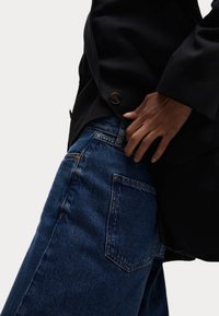 Blazer oscuro con detalle de botones, combinado con jeans de denim azul que presentan un diseño clásico de cinco bolsillos y sutiles acentos de costura.