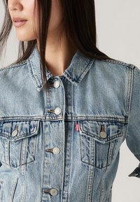 Chaqueta de mezclilla azul claro con dos bolsillos en el pecho, botones plateados y un cuello clásico. Textura desgastada y detalles de costura visibles.