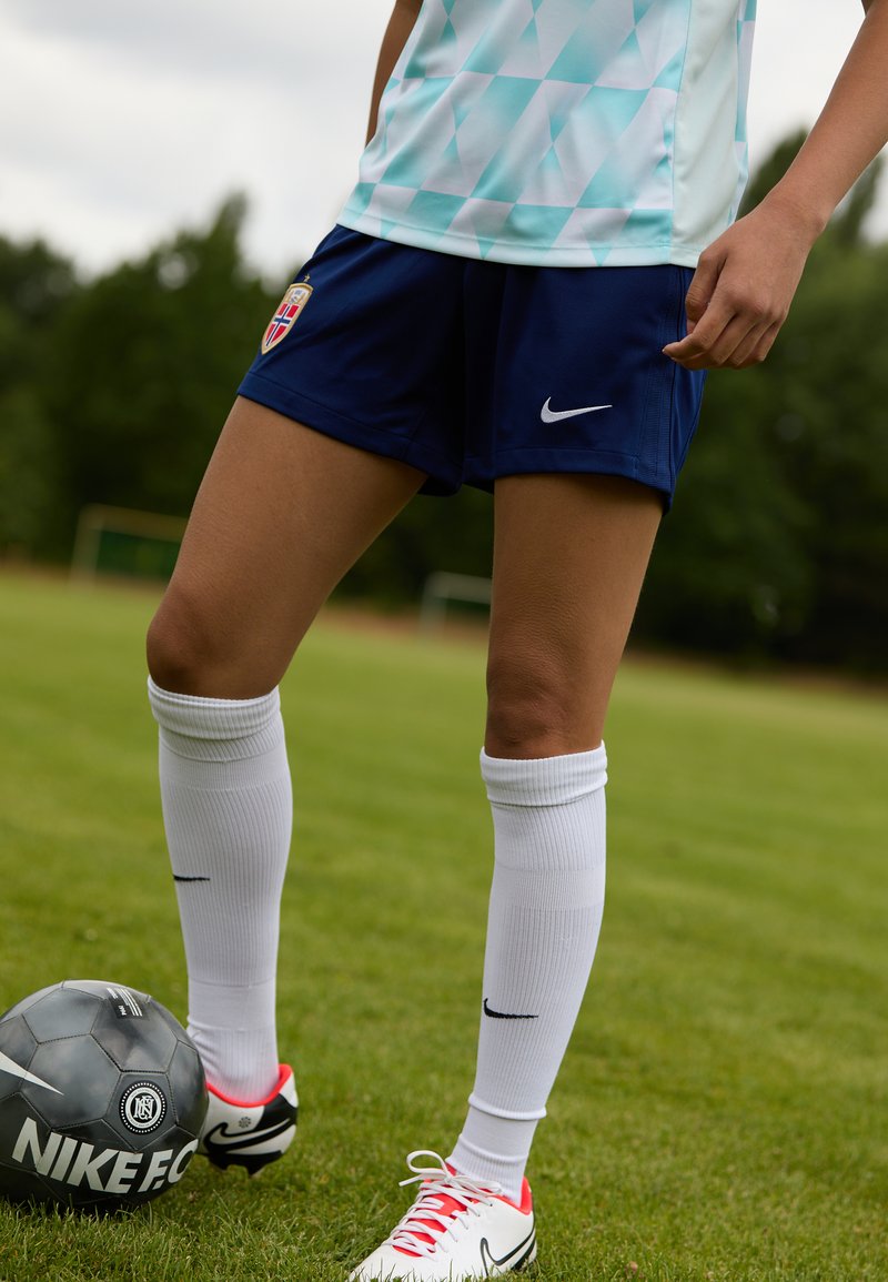 Blå sportshorts med Nike-logga, ljus turkos skjorta med geometriskt mönster, vita strumpor och fotbollsskor med röda detaljer bredvid en svart Nike-boll.