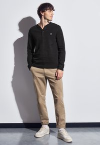 Man i svart långärmad henley-tröja, beige byxor och vita sneakers som står mot en vit vägg, tittar åt höger med händerna delvis i fickorna.