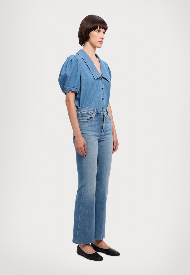 Denim-paitta, jossa on ylisuuri kaulus, napit edessä ja puffat hihat. Yhdistetty vaaleat farkut, joissa on leveät lahkeet, ja mustat ballerinat. Yksinkertainen tausta.