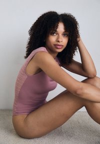 Fitted bodysuit in soft pink fabric with a textured mesh panel on the side; sleeveless design and smooth finish. Model sitting on the floor.