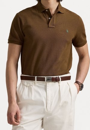 Mann trägt braunes Poloshirt, weiße plissierte Hose, braunen Ledergürtel und Armbanduhr mit braunem Band, Hände in den Taschen.
