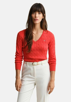 Kvinde med langt brunt hår iført en rød kabelstrikket V-hals sweater og hvide højttaljede bukser med bælte, stående foran en ensfarvet baggrund.