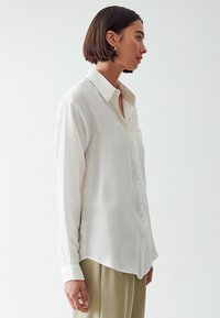 Blusa bianca a maniche lunghe in un tessuto leggero e trasparente, con chiusura a bottoni davanti, colletto e una vestibilità comoda. L'orlo è leggermente curvato ai lati.