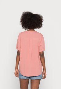 Helle rosa Bluse mit kurzen, hochgerollten Ärmeln, einer Rückenfalte und einem abgerundeten Saum. Getragen mit blauen Jeansshorts. Glatte Stofftextur.