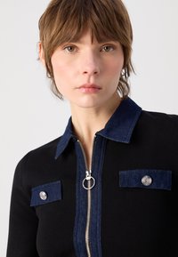 Haut zippé noir avec un col en denim bleu foncé et des accents de poches. La texture est lisse avec une fermeture éclair en métal et des détails de boutons décoratifs.