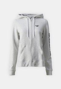Sudadera gris con cremallera, hecha de algodón suave. Cuenta con capucha con cordón, bolsillos laterales y logo negro en el pecho y la manga.