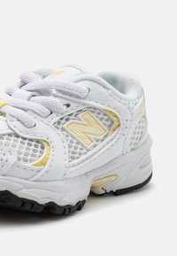 Balti tinklinio bateliai su geltonais akcentais, apvalia forma, tekstūruota padas ir storos raišteliai. Šone yra logotipas ir sustiprinta kulno dalis.