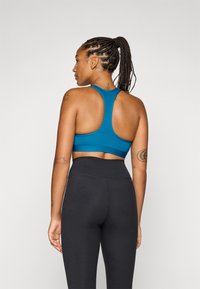 Una persona con capelli intrecciati indossa un top atletico blu e leggings neri, rivolta di spalle su uno sfondo grigio chiaro.