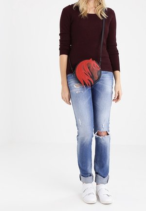 Rote und schwarze kreisförmige Umhängetasche mit Pferdekopf-Design, ausgestattet mit einem langen Riemen und glattem Material, getragen zu lässigen Jeans und weißen Schuhen.