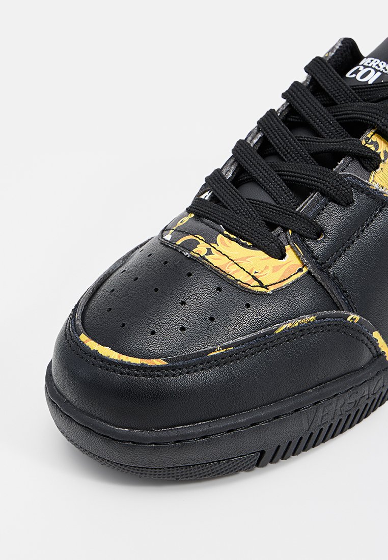 Zapatilla negra con parte superior de cuero suave y acentos florales amarillos, cordones texturizados negros, puntera perforada y suela de goma.