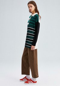 Zaļš un melns svītrots garām piedurknēm polo krekls ar baltu apkakli, valkāts ar brūniem platām biksēm un sarkaniem sporta apaviem ar baltām akcentiem.