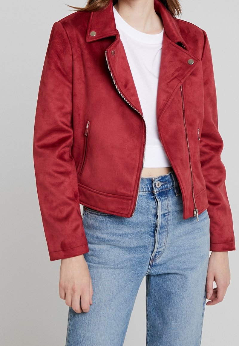 Mujer con una chaqueta biker de ante roja sobre un top corto blanco y unos vaqueros de talle alto azul claro sobre un fondo gris liso.