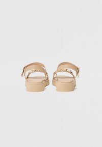 Beige sandaler med en tykk, teksturert såle, ankelrem og gullfarget detaljer. Enkelt, minimalistisk design med justerbar spenne.