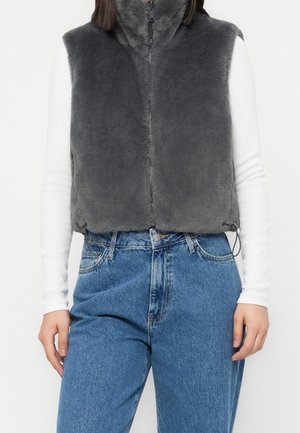 Person trägt eine ärmellose dunkelgraue flauschige Zip-Up-Weste über einem langärmligen weißen Oberteil, kombiniert mit hoch taillierten blauen Jeans.