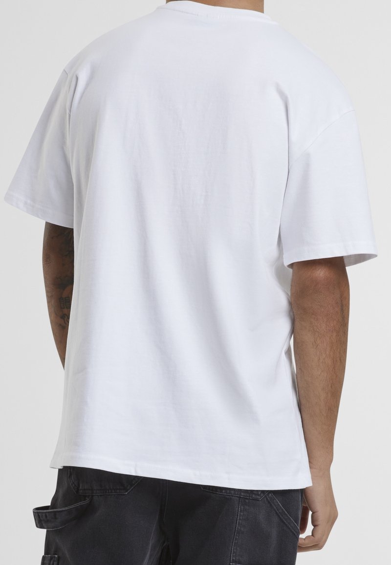 Homme portant un T-shirt blanc uni à manches courtes et un pantalon cargo noir, vu de dos sur un fond neutre.