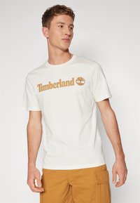 Fehér pamut póló barna "Timberland" logóval, rövid ujjú és kerek nyakrészű kivitelben. Bézs cargo sorttal párosítva.
