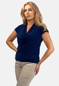 Granatowa bluzka z drapowaniem i krótkimi rękawami, z dekoltem typu cowl. Gładka tekstura, noszona z jasnobeżowymi dopasowanymi spodniami. Casualowy styl.