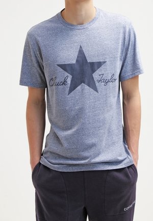 T-shirt en coton gris avec un grand motif d'étoile sombre et le texte « Chuck Taylor » dans une police en écriture. Manches courtes et encolure ras du cou.