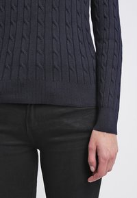 Pull bleu marine en maille torsadée avec ourlet côtelé porté avec un pantalon noir, montrant un gros plan du torse et de la main droite.