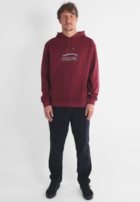 Haut à capuche bordeaux avec poche avant et cordon de serrage, comportant un graphique "OXBOW" en bleu et blanc. Assorti avec un pantalon sombre et des chaussures noires.