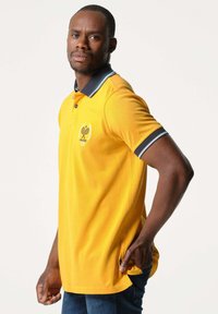 Geel polo shirt met een marineblauwe kraag en manchetten, met een tennislogo op de borst. Gemaakt van lichte stof, met een rechte pasvorm.