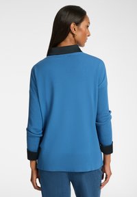 Frau, die mit dem Rücken zur Kamera steht, trägt einen blauen Langarmpullover mit schwarzem Kragen und Bündchen, kombiniert mit blauen Hosen, vor einem weißen Hintergrund.