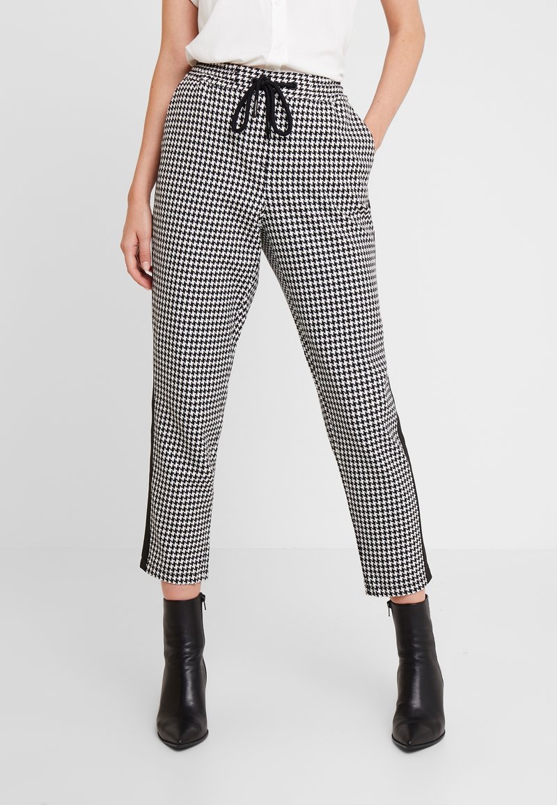 Kalhoty s klasickým houndstooth vzorem v černobílé barvě, s elastickým pasem, stahovací šňůrkou a bočními pruhy, spárované s černými kotníkovými botami.