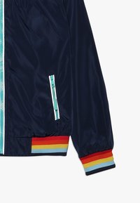 Navy blue jacket sleeve with teal zipper, side pocket zipper, and ribbed cuff featuring red, yellow, light blue, and navy stripes.