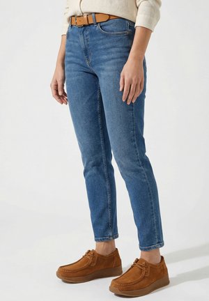 Person trägt einen hellbeigen Pullover, blaue Jeans mit braunem Gürtel und braune Wildleder-Schnürschuhe und steht vor einem schlichten Hintergrund.