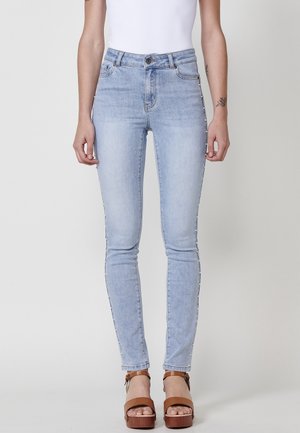 Vrouw draagt lichtblauwe skinny jeans met witte parels langs de buitennaad en bruine plateauzandalen, staand tegen een effen witte achtergrond.