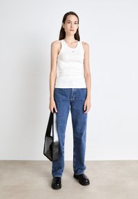 White ribbed tank top with a logo, paired with blue straight-leg jeans, black platform shoes, and a black crossbody bag. Plain background.