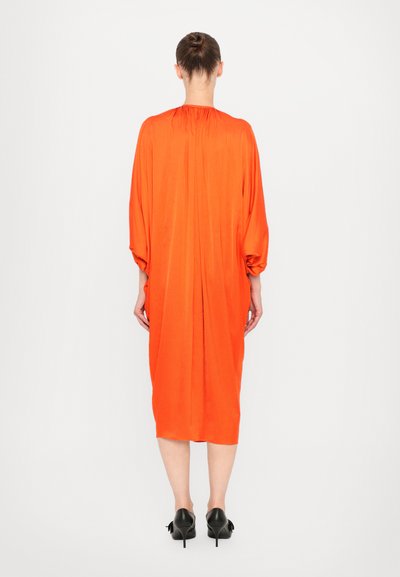 Vestido naranja brillante con un diseño suelto y fluido. Mangas largas con fruncidos en los hombros y dobladillo a la altura de la rodilla. Combinado con zapatos negros de tacón.