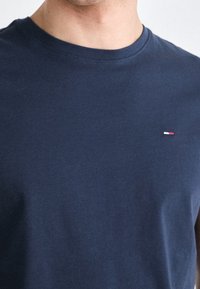 Námořnická modrá bavlněná tričko s krátkým rukávem, které má kulatý výstřih a malou logo akcentaci v červené, bílé a modré barvě na levé části hrudi.